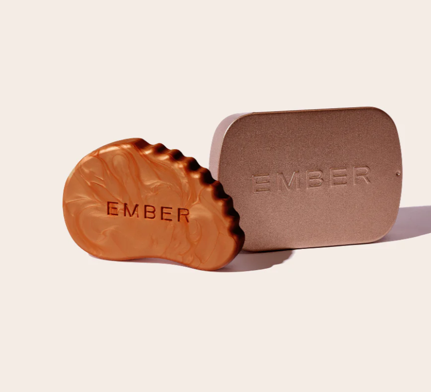 A bronze skincare highlighting bar shaped like a gua sha skincare tool is sitting beside a metallic tin with the word "Ember" embossed on it.