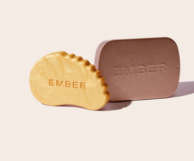 A solid skincare highlighting bar called Sunstone which is shaped like a gua sha tool sits beside a metallic tin with the word "Ember" embossed on it. 