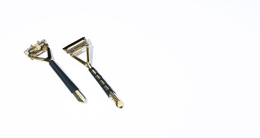 Two gold razors lay on a white surface with a black grip sleeve on the handle.