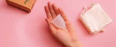Image of two hands holding red menstrual cups, the background is a soft pink colour.