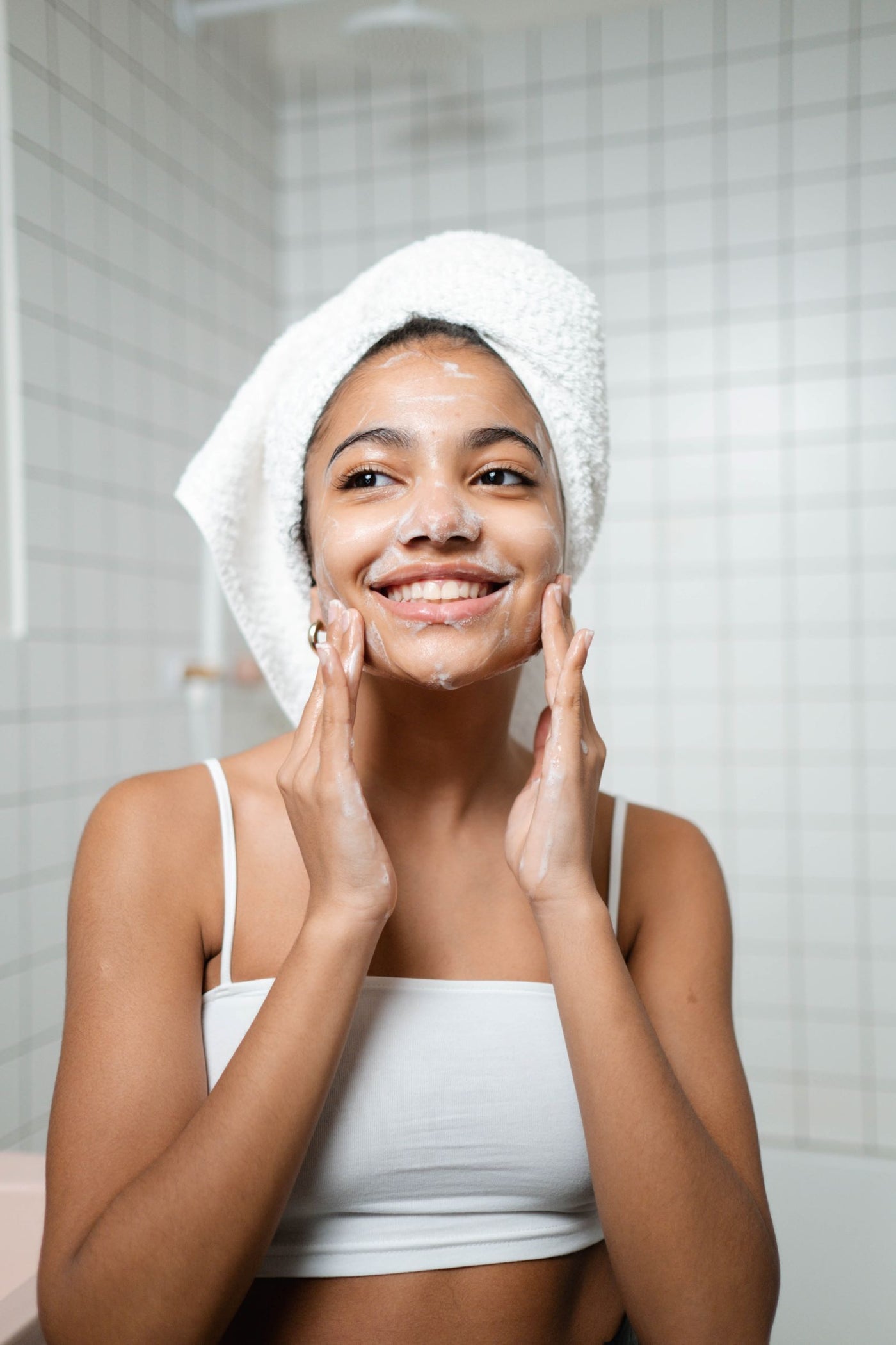 Image of a young, brown skinned girl with a white towel on her head applying a facial wash on her face. 