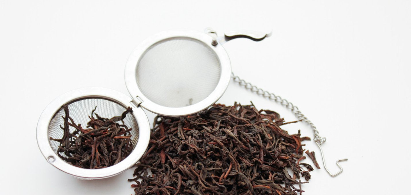 Stainless steel clamp strainer on a checkered tea towel, beside a white bowl filled with loose leaf tea. 