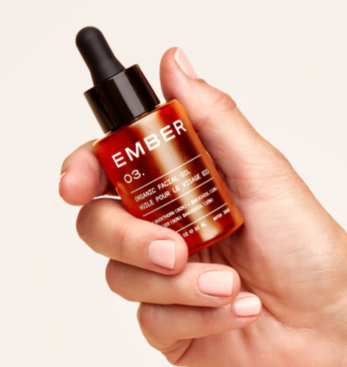 A hand holding a 12 ml facial oil in a glass amber bottle with a black dropper top. 