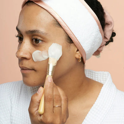 Image of a person with their hair tied back with a white and pink headband, using mask applicator with wooden handle to apply a white face mask to their cheek
