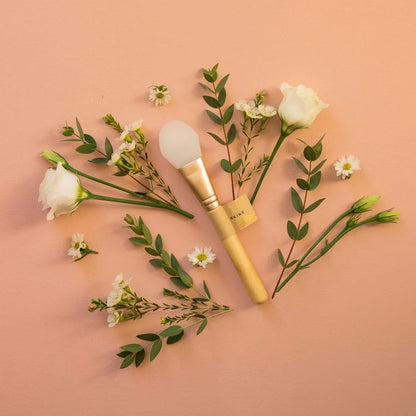 Image contains mask applicator on a pinkish background, surrounded by leaves and flowers