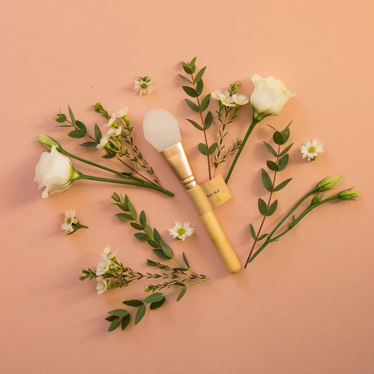 Image contains mask applicator on a pinkish background, surrounded by leaves and flowers