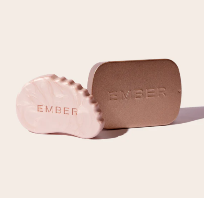 A skincare highlighting bar called Rose Quartz which is shaped like a gua sha skincare tool sits beside a metallic tin with the word "Ember" embossed on it. 