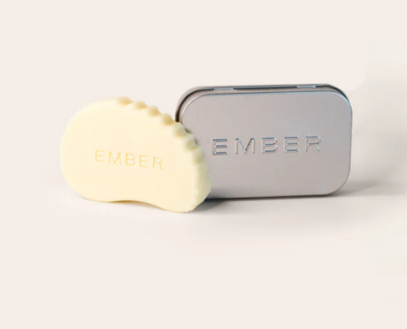 A skincare serum bar shaped like a gua sha skincare tool is placed beside a metal tin with the words "Ember" embossed on it. 