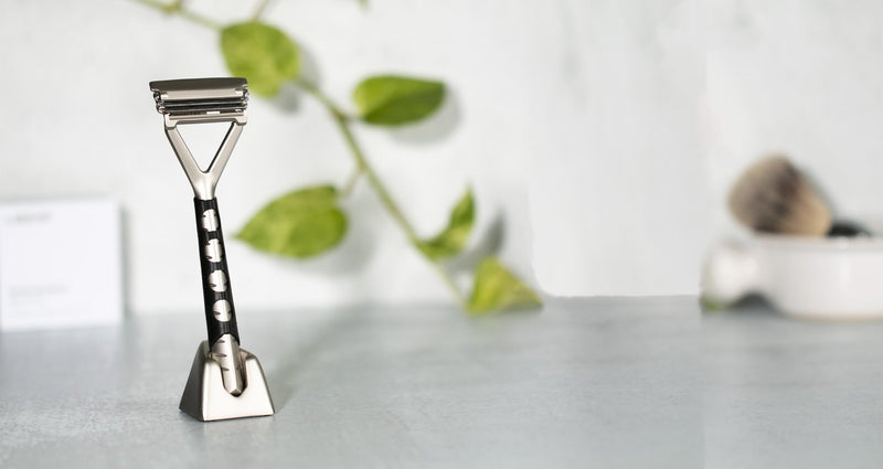 A chrome razor sits in its stand on a counter top. The razor has a black grip sleeve on the handle of the razor. There is a plant vine in the background.