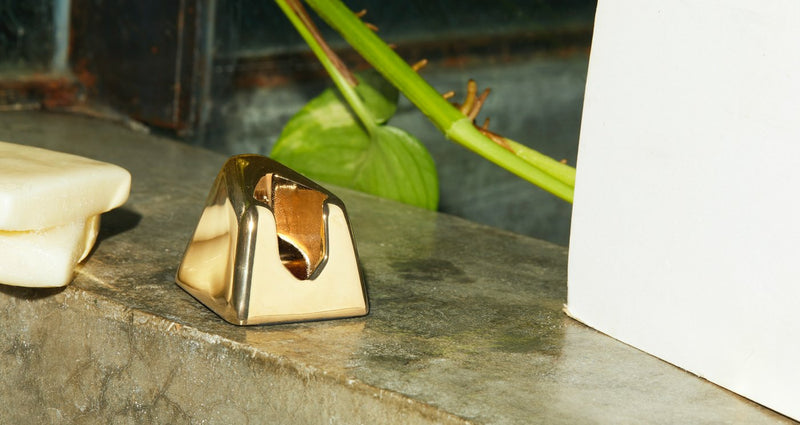 A gold razor stand sits on a counter with a plant in the background